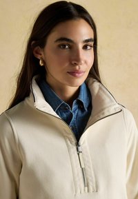 Kremowy polar z półzapięciem i wysokim kołnierzem, z dżinsową koszulą pod spodem oraz metalowym suwakiem. Gładka faktura.