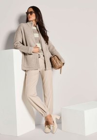 Eine Frau trägt eine beige Strickjacke, einen gestreiften Pullover, beige Jogginghosen und cremefarbene Blockabsatzslipper und hält eine gequiltete braune Handtasche.