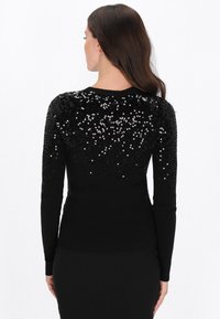 Fekete pulóver hosszú ujjakkal, flitterekkel a háton, bordázott textúrával és testhez simuló fazonnal. Egyszerű kerek nyakkal.