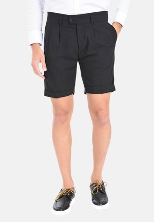 Pantaloni corti neri su misura con pieghe, risvolti e tasche laterali. Abbinati a scarpe nere con suole bianche e lacci gialli.