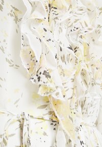 Květinové šaty s volánkovými detaily, vyrobené z jemné bílé látky zdobené vzory žlutých a zelených květin, doplněné jemným šitím.