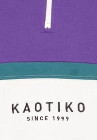 Pullover viola con cerniera bianca, caratterizzato da blocchi di colore turchese e bianco. Logo nero ricamato con la scritta "KAOTIKO SINCE 1999." Texture liscia.