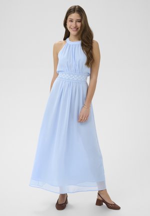 Naeratav naine, kes kannab pikka helerohelist läbipaistmatut kleiti pitsdetailiga vöökohal ja pruunide kontsakingadega, seisab valgel taustal.