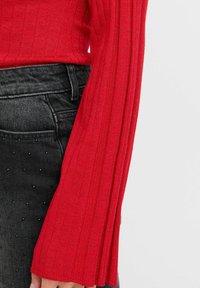 Röd ribbad tröja med långa klockärmar, texturerat tyg och en figurnära design; matchad med mörkgrå denim som har dekorativa nitar.
