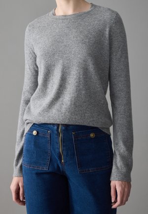 Personne portant un pull gris à manches longues et un jean bleu taille haute avec de grandes poches avant et une fermeture éclair visible.