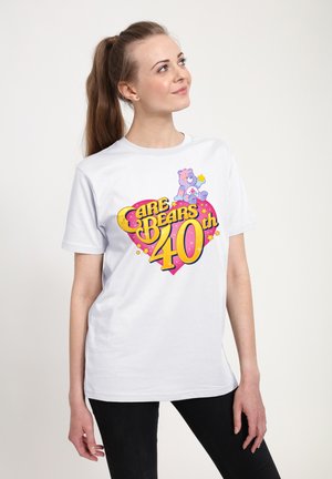 Hvid bomulds t-shirt med et farverigt grafisk design, der har teksten "Care Bears 40th" og en bjørneillustration på en pink hjertebaggrund.