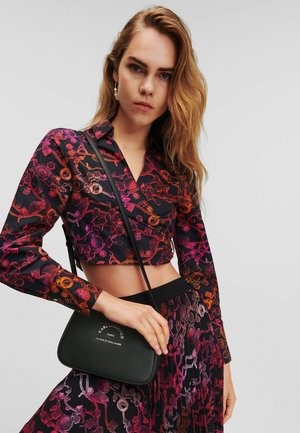 Bolso bandolera negro con un acabado texturizado, que presenta un estampado de logo circular en blanco, sostenido por un modelo que lleva una camiseta corta con estampado.