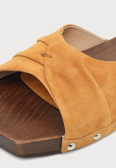 Gros plan d'une sandale en daim couleur fauve avec de larges brides superposées, une semelle en bois et des clous en métal argenté sur le bord.