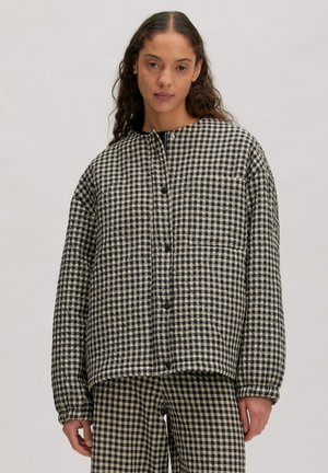 Frau trägt schwarz-beige karierte gesteppte Jacke und passende Hose, steht vor einem schlichten hellen Hintergrund.