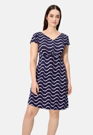Mujer con un vestido azul marino con patrones de olas blancas, mangas cortas con volante, escote en V y detalle de lazo delantero, posando frente a un fondo liso.