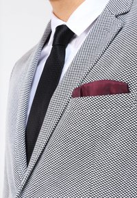 Man som bär en svart strukturerad slips, vit skjorta och en grå mönstrad blazer med en vinröd fickfyllning.