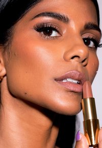 Nude läppstift hålls nära läpparna, gyllene metallhölje, slät textur, kombinerat med väl definierade ögonfransar, naturlig sminkning och brun hudton.