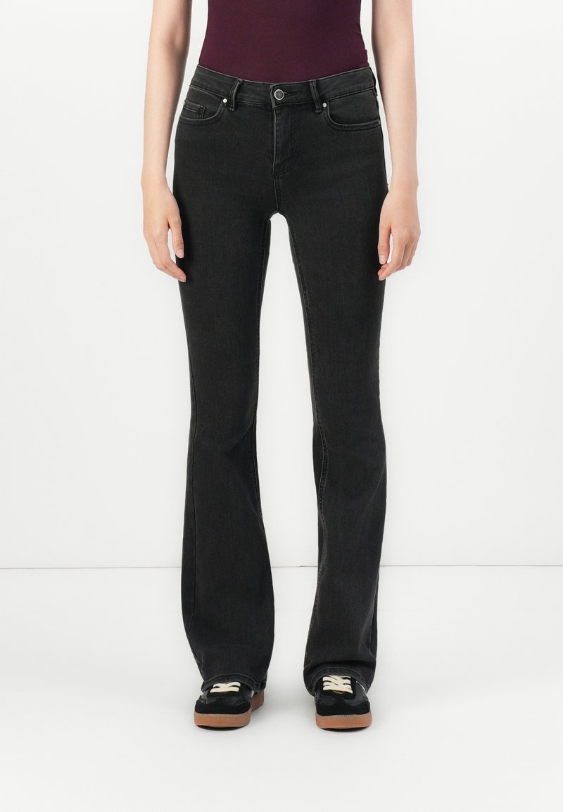 Svarte buksene med flared design laget av denim, med en tettsittende midje, to frontlommer og knappelukking. Sammen med svarte joggesko.