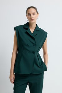 Žena s světlou pletí a staženými vlasy, oblečená v tmavozeleném bezrukávovém dvouřadém blejzru a odpovídajících kalhotách na bílém pozadí.
