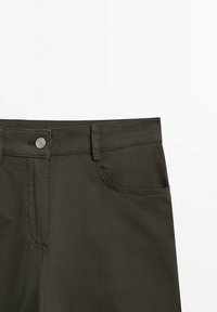 Gros plan sur un pantalon marron foncé avec un bouton en métal, des passants pour ceinture et une poche avant, présenté sur un fond blanc uni.