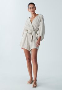 Romper w jasnym beżu z pionowymi paskami, z dużymi rękawami, paskiem w talii i krótkimi spodniami. Gładka tekstura, minimalistyczny design.