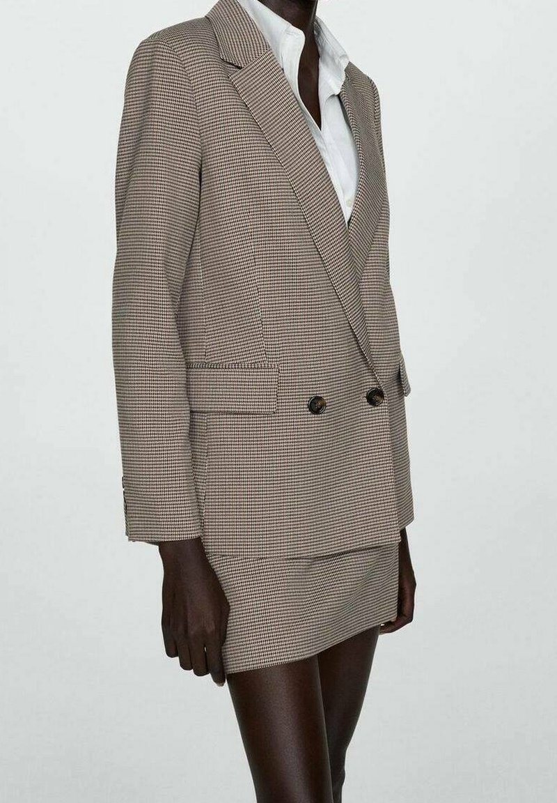 Femme portant un blazer croisé beige et marron à carreaux avec une jupe assortie et une chemise blanche sur un fond uni.