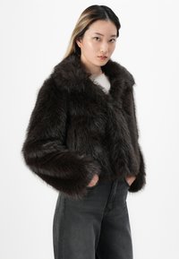 Chaqueta recortada de piel sintética marrón con un amplio collar, que cuenta con mangas largas y una textura suave. Lleva puesta sobre una blusa clara y jeans oscuros.