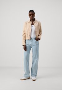 Jachetă bej scurtă, top alb din bumbac, blugi deschiși la culoare cu talie înaltă și adidași albi. Ochelari de soare cu nuanță maro, design minimalist.