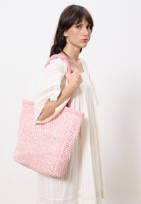 Rosa gewebte Tragetasche mit strukturierten, handgefertigten Details, ausgestattet mit stabilen Trägern und einem geräumigen Innenraum, gehalten von einer Person in einem hellen Kleid.