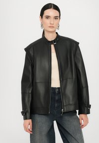Jachetă din piele neagră cu fermoar în față, umeri structurați și curele la manșete, purtată peste un top bej, asortată cu pantaloni din denim închis, lejeri.