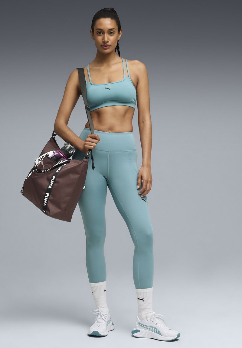 Atletisk kvinde iført turkis sports-bh og leggings, hvide Puma-sneakers, holder en brun Puma træningstaske med en vandflaske på skulderen.