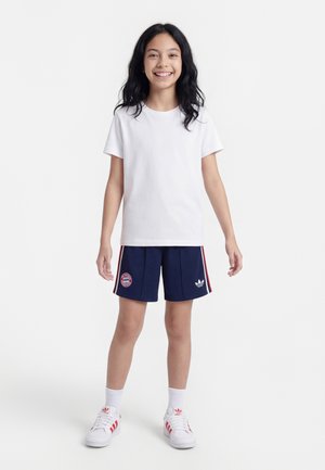 Smilende barn med langt sort hår iført en hvid t-shirt, marineblå sportsshorts med logoer, hvide sokker og hvide sneakers med røde striber.
