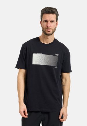 Uomo che indossa una t-shirt nera Armani Exchange con grafica a righe orizzontali bianche e pantaloni neri, in piedi davanti a uno sfondo chiaro uniforme.