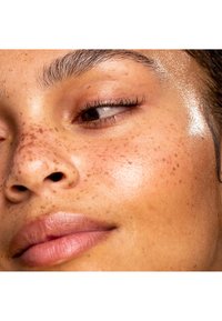 Gros plan sur un visage avec un teint lisse et légèrement tacheté. La peau semble légèrement brillante. Les sourcils sont bien entretenus et les lèvres sont d'un rose naturel.