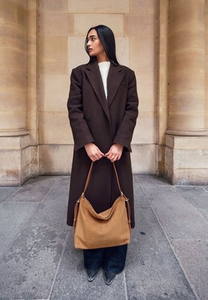 Abrigo de lana marrón, suéter crema, jeans oscuros, botas puntiagudas grises. Lleva una bolsa de ante marrón con una forma estructurada y acentos de cuero.