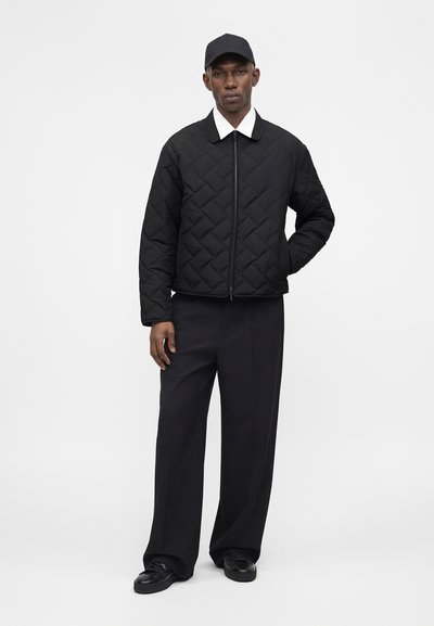 Άνδρας με μαύρο καπιτονέ μπουφάν με φερμουάρ, μαύρο φαρδύ παντελόνι, μαύρο καπέλο και μαύρα παπούτσια, στέκεται με το ένα χέρι στην τσέπη.