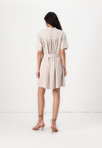 Beige linen dress with short sleeves, a cinched waist, and a back pleat detail, featuring a smooth texture and knee-length cut.