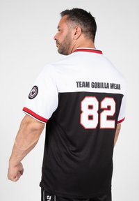Camisa deportiva en blanco y negro con acentos rojos, que presenta "TEAM GORILLA WEAR" y el número "82" en la parte trasera. Manga corta.