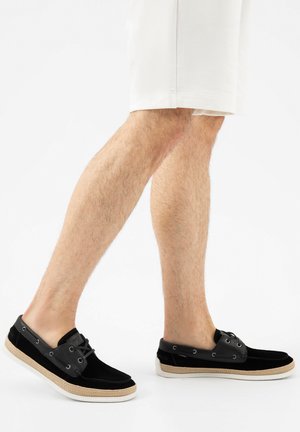 Beine in weißen Shorts und schwarzen Bootsschuhen mit beigen Sohlen, die vor einem einfachen weißen Hintergrund gehen.