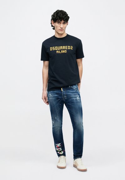 Marineblå bomulds-T-shirt med gul "DSQUARED2 MILANO"-tryk, kombineret med slidte blå jeans med multifarvede accenter og beige sneakers.