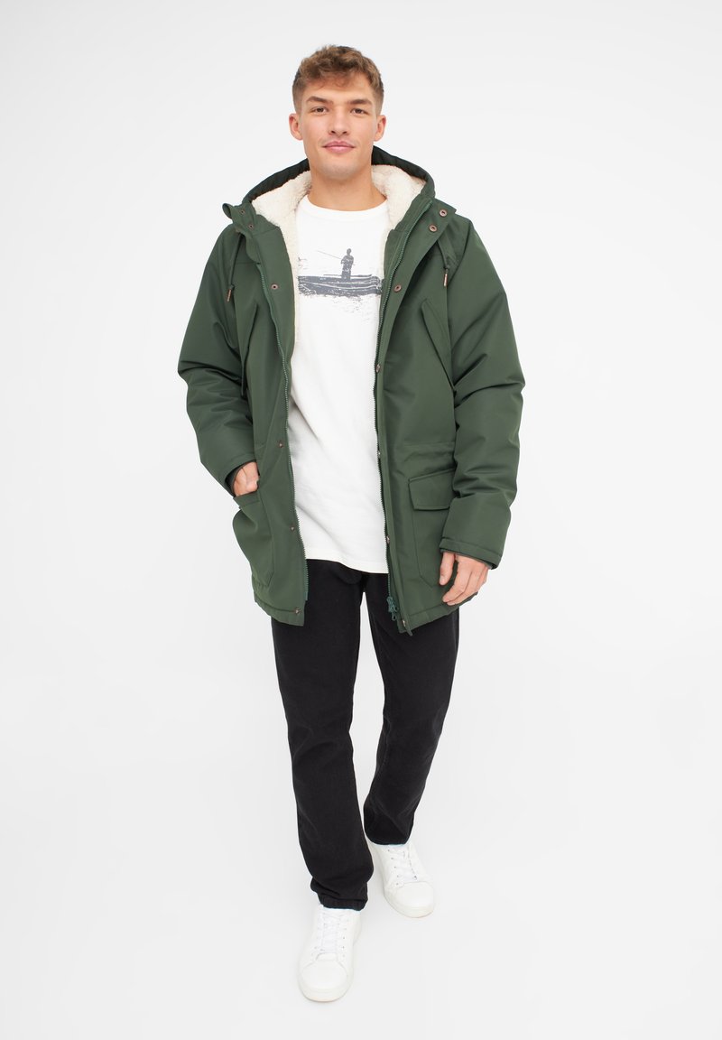 Parka impermeabile verde con cappuccio foderato in pelliccia, chiusura a zip e bottoni a pressione, tasche laterali, indossata sopra una t-shirt bianca con grafica e pantaloni neri.