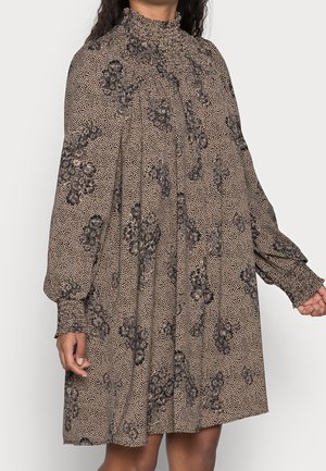 Kvinde iført beige minikjole med lange ærmer, sort blomsterprint, høj rynket krave og smockede manchetter.