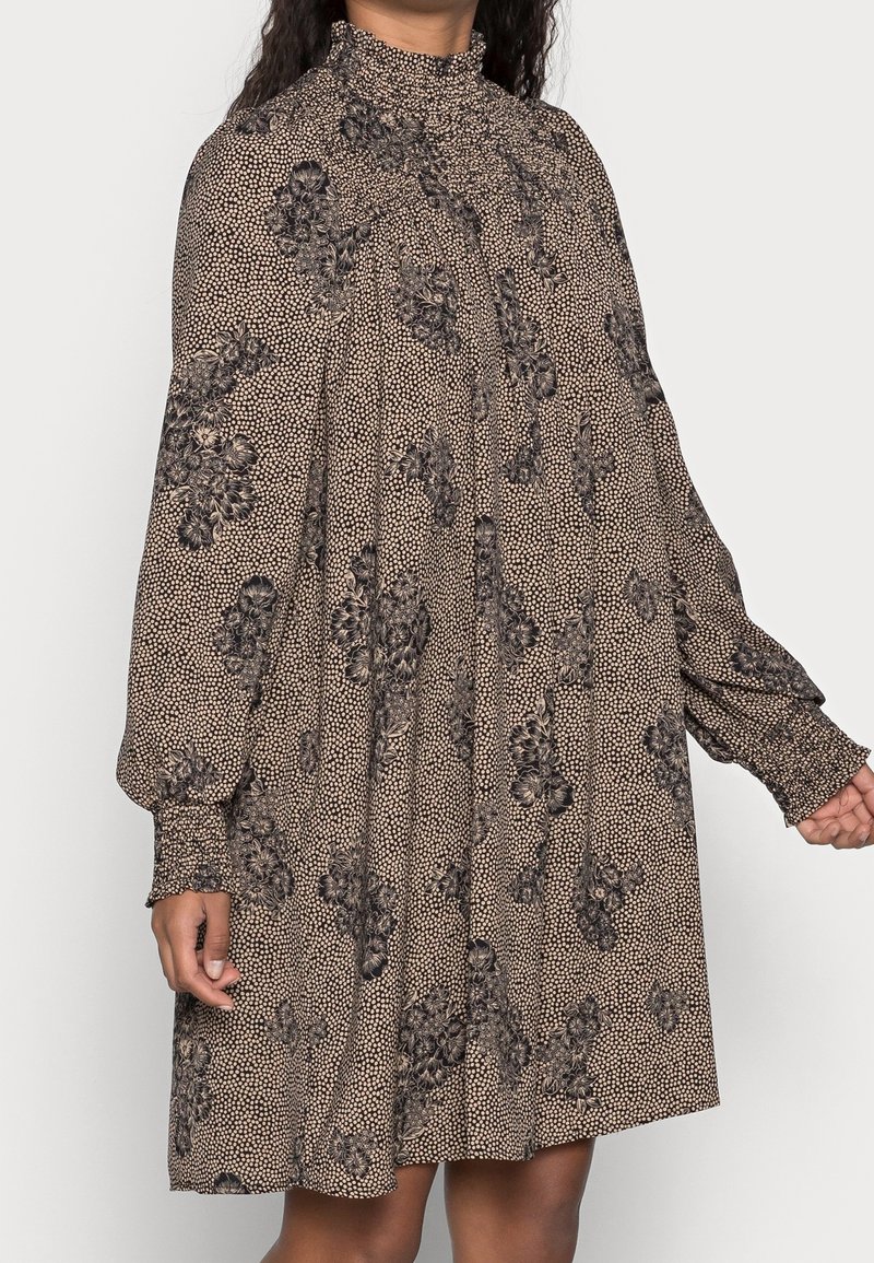 Femme portant une mini-robe beige à manches longues avec imprimé floral noir, col montant froncé et poignets smockés.