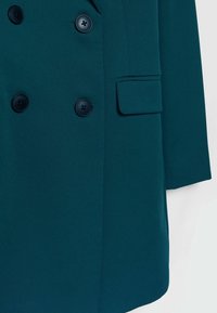 Manteau croisé bleu sarcelle avec des boutons noirs et une poche à rabat sur le côté droit.