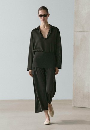 Woman walking indoors wearing loose black top and wide black pants, dark sunglasses, and beige ballet flats.