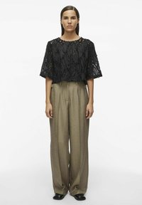 Top corto negro texturizado con patrón de olas, combinado con pantalones sueltos de talle alto en tono topo. La modelo está de pie frente a un fondo neutro.