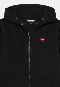 Sweatshirt preto com fecho, com uma textura suave, apresentando um capuz com cordão, bolsos tipo canguru e um pequeno logótipo vermelho da Wrangler no peito.