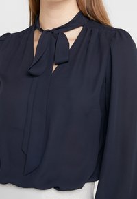 Námořnická modrá blůza s dlouhými puff rukávy a zavazováním u výstřihu. Měkký materiál s mírným leskem a výřezem na ramenech.