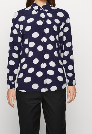 Mujer con camisa de manga larga azul marino con grandes lunares blancos y pantalones negros, de pie contra un fondo liso.