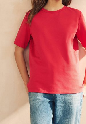 Woman wearing a plain red short-sleeve t-shirt and light blue jeans, standing with hands resting at her sides.