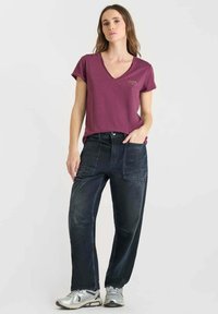 Vrouw in een paarse V-hals T-shirt, zwarte relaxte patch pocket broek en zilveren sneakers, staande tegen een lichte effen achtergrond.