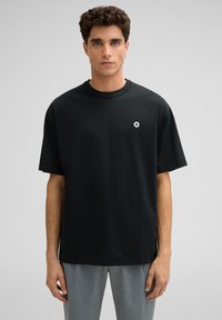 Junger Mann mit kurzen, lockigen Haaren, der ein lockeres schwarzes T-Shirt mit einem kleinen weißen Kreuzlogo auf der Brust und grauen Hosen trägt, steht.