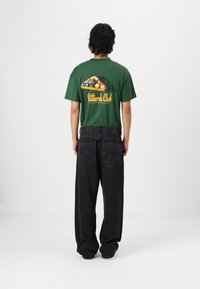 Tricou din bumbac verde cu un grafic de biliard pe spate, asociat cu pantaloni din denim negri, largi. Designul include litere îndrăznețe și culori vibrante.