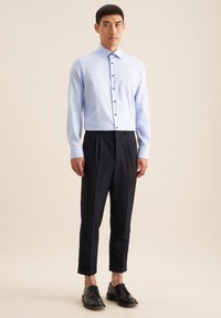 Camicia a maniche lunghe azzurra chiara con colletto e bottoni scuri, abbinata a pantaloni neri a sbuffo e scarpe nere con lacci.