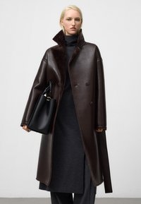 Cappotto in pelle marrone con colletto alto e chiusura a bottoni, dotato di un interno morbido e spacchi laterali. Abbinato a una borsa nera.
